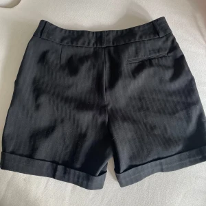 Svarta dressade shorts med uppvik - Klassiska svarta shorts med dressad look och uppvikta benslut. De har en rak passform, diskreta fickor och bälteshällor i midjan. Perfekta för en clean och stilren outfit. Materialet känns mjukt och svalt, troligen bomull eller polyester Märket Caroll köpt i Paris 2004 