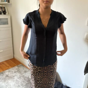 Svart blus med volangärm - Svart blus med korta volangärmar och v-ringning. Framsidan har snygga veckade detaljer som ger en stilren look. Blusen är lätt och luftig, perfekt till både jeans och kjol. Materialet känns mjukt och följsamt mot huden.