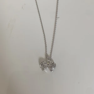Silverhalsband med glittrig rosett - Supersött halsband i silverfärg med ett hänge format som en rosett, täckt av gnistrande stenar. Kedjan är tunn och smidig, perfekt för att ge en extra touch till din outfit. Rosetten ger ett gulligt och trendigt intryck.