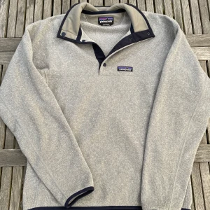 Grå fleece-tröja från Patagonia - Säljer en grå fleece-tröja från Patagonia med mörkblå detaljer och hög krage. Tröjan har knappar vid halsen och är långärmad. Perfekt för kyliga dagar och riktigt skön att ha på sig. Patagonia-logga på bröstet.