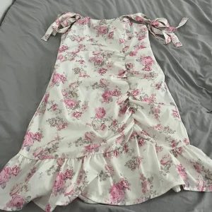 Vit blommig kortklänning med volang - Supersöt vit kortklänning med rosa och beige blommor, knytbara axelband och volangkant nertill. Klänningen har rynkade detaljer framtill och är perfekt för sommarens alla tillfällen. Lätt och luftig känsla, passar dig som gillar romantisk stil.