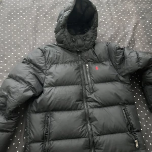 Svart dunjacka från Polo Ralph Lauren - Svart dunjacka från Polo Ralph Lauren med huva och dragkedjor framtill. Jackan har en klassisk pufferstil och är dekorerad med den ikoniska röda loggan på bröstet. Perfekt för kalla dagar och riktigt snygg till streetwear. Priset går absolut att diskuteras vid snabb affär