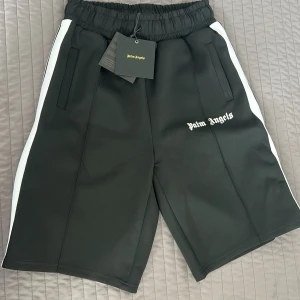 Palm Angels svarta shorts med vita ränder - Svarta shorts från Palm Angels med vita sidoränder och broderad logga framtill. De har elastisk midja och två fickor. Materialet är en mix av bomull och polyester, vilket ger en sportig och skön känsla. Dem är helt oanvända bara testade taget är fortfarande kvar 