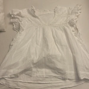 Vit blus med volang från Gina Tricot - Supersöt vit blus från Gina Tricot med volangdetaljer på ärmarna och v-ringning. Lätt och luftig i bomull, perfekt för varma dagar. Blusen har en romantisk känsla med rynkade partier och korta ärmar.
