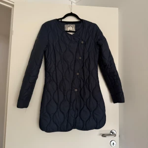 Quiltad svart kappa från myMO - Snygg svart quiltad kappa från myMO med lång passform och rund halsringning. Jackan har assymetrisk knäppning med knappar framtill och ett stilrent mönster i tyget. Perfekt för dig som vill ha en modern och enkel look.