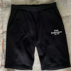 Svarta shorts Peak Performance 170 - Svarta shorts från Peak Performance med broderad vit logga på ena benet. Modellen har elastisk midja och två sidofickor. Perfekta för träning eller chill, klassisk och stilren look. Materialet känns mjukt och sportigt.