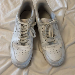 Nike Air Force 1 vita sneakers - Klassiska Nike Air Force 1 sneakers i helvitt skinn med perforerad tå och vit sula. Skorna har rund tå, snörning och diskret Nike-logga på sidan. Perfekta för en clean och tidlös streetstil.