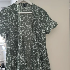 Grön prickig omlottklänning - Supersöt grön kortklänning från Vero Moda  med vita prickar. Klänningen har korta ärmar, lätt v-ringning och volangkant nertill. Perfekt flowy passform och skön att bära under varma dagar. Materialet känns lätt och luftigt.