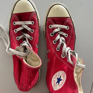 Röda Converse Chuck Taylor All Star - Klassiska röda Converse Chuck Taylor All Star high tops med vit sula och tå, vita snören och tydliga kontrastsömmar. Skorna har rund tå, platt sula och ikonisk logga på sidan. Ovandel i textil och detaljer i vitt och blått. Storlek 37,5. 