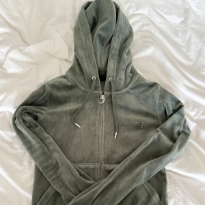 Mörk grön hoodie Juicy Couture  - Mjuk och lyxig grå hoodie från Juicy Couture i velour med dragkedja, snörning i huvan och broderad JC-logga på bröstet. Perfekt för en chill och trendig look. Långärmad och med klassisk passform.