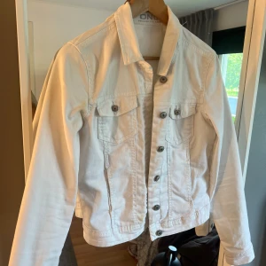 Vit jeansjacka från ONLY, strl 38 - Snygg vit jeansjacka från ONLY i klassisk modell med knappar framtill och två bröstfickor. Jackan har lång ärm och är tillverkad i bomull, perfekt för vår och sommar. Enkel att matcha med allt och ger en fräsch look.