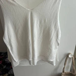 Vit ribbad linnetopp med v-ringning - Säljer en stilren vit linnetopp med v-ringning och ribbad struktur. Toppen är ärmlös och har en avslappnad passform, perfekt för varma dagar. Materialet känns lätt och luftigt, och den har en snygg, enkel design som är lätt att matcha.