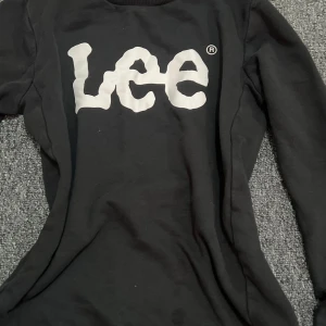 Svart långärmad tröja från Lee - Svart långärmad tröja från Lee med stor vit logga framtill. Klassisk rund halsringning och mjukt material som känns skönt mot huden. Perfekt för dig som gillar enkel och stilren streetwear med tydlig branding.
