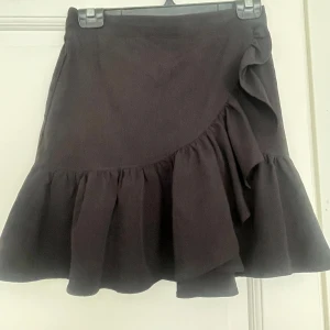 Svart volangkjol från Lindex - Supersöt svart kortkjol från Lindex med volangdetaljer och omlottkänsla framtill. Kjolen har en elastisk midja som gör den bekväm och lätt att bära. Perfekt att styla med en crop top eller stickad tröja för en trendig look.