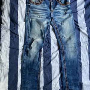 Blå jeans från Cipo & Baxx med broderi - Säljer ett par blå jeans från Cipo & Baxx med snygga slitningar och hål på låret. Jeansen har coola broderade bakfickor i brunt och kontrastsömmar. Klassisk femficksmodell med normal passform och raka ben. Perfekta för dig som gillar streetstyle.