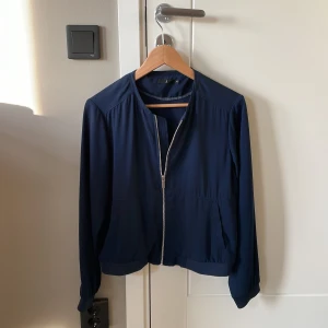 Mörkblå bomberjacka med dragkedja - Snygg mörkblå bomberjacka med silvrig dragkedja framtill och två fickor. Jackan har en clean look utan krage, långa ärmar och elastisk nederkant. Perfekt för dig som gillar stilrena och enkla plagg. Passar till det mesta och är lätt att matcha. Det är ett väldigt tunt material som kan passa som både jacka eller tröja över ex ett linne. Jackan är från Åhléns, st 40. Jag har alltid S i kläder och den passar mig väldigt bra så den är väldigt liten i storleken!! 💙