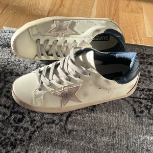 Golden Goose Super Stars | 39 - Golden Goose sneakers i vitt skinn med beige mockastjärna på sidan och blå metallic-detalj vid hälen. Klassisk rund tå, platt sula och grå snörning. Snygg kontrast mellan material och färger, perfekt för en trendig streetstyle-look. Storlek 39