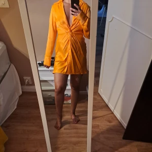 Kort orange omlottklänning - Säljer en kort, orange klänning med omlott-effekt och djup v-ringning. Klänningen har långa ärmar, knappar framtill och snygg drapering i midjan. Perfekt för dig som vill sticka ut med en färgstark look! Endast använd en gång.