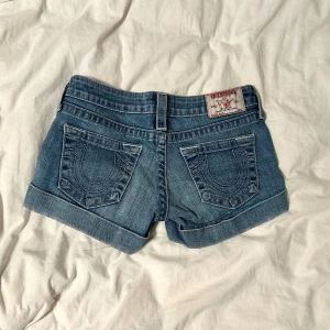 True Religion jeansshorts blå, strl 26 - Snygga blå jeansshorts från True Religion med klassiska bakfickor och uppvikta benslut. Shortsen har silverfärgade knappar och detaljer, samt bälteshällor. Perfekta för sommaren och ger en cool, avslappnad vibe.