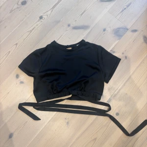 Svart croppad topp med knytband - Svart croppad topp med korta ärmar och långa knytband i midjan. Enkel och stilren design som passar perfekt till höga jeans eller kjol. Mjuk och lätt i materialet för en bekväm känsla.