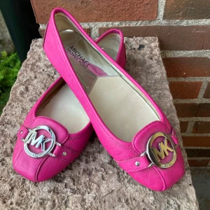 Rosa ballerinaskor Michael Kors stl 39 - Snygga rosa ballerinaskor från Michael Kors med rund tå och ikoniskt MK-spänne i silver framtill. Skorna är i skinnimitation med gummisula och har en stilren design som sticker ut. Perfekta för dig som vill ha en poppig färg och lyxig känsla. Storlek 39. Kan även passa 38.5. 