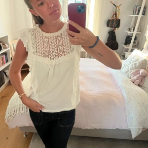 Vit blus med spets och volang - Superfin vit blus med broderad spets upptill och volangdetaljer längs axlarna. Ärmlös modell med rund halsringning och romantiska broderier. Perfekt för dig som gillar en söt och somrig vibe. Tillverkad i mjuk bomull och knapp i nacken. 