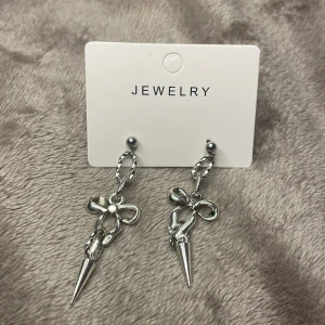 Silvriga örhängen med rosett och spike - Snygga silvriga örhängen med coola detaljer! Hängande design med tvinnad oval länk, en metallisk rosett och en spetsig spike längst ner. Perfekt accessoar för dig som vill sticka ut och addera lite edge till din stil.