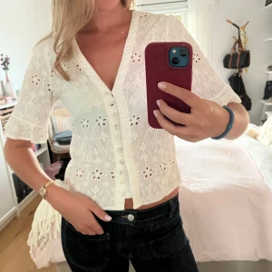Vit broderad blus med knappar - Superfin vit blus med broderade blommor och hålmönster. Kort ärm med lätt volangkant, v-ringning och pärlemorknappar framtill. Croppad modell som ger en fräsch och somrig vibe. En knapp ”hylsa” som gått sönder men syns inte nät blusen är knäppt och borde vara lätt att laga om man vill det 💛