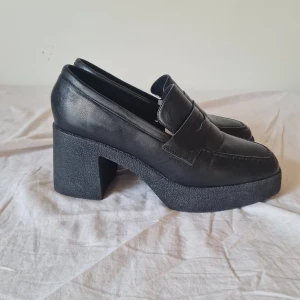 Svarta chunky loafers med klack - Snygga svarta loafers med chunky hög klack och plattformsula. Skorna har klassisk penny loafer-design med markerad söm och är tillverkade i skinnimitation. Perfekta för dig som vill ha en edgy och trendig look. Originellt pris: 800kr. Använt 1 gång