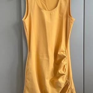 Gul ärmlös kortklänning från Sisley - Snygg gul ärmlös kortklänning från Sisley med rund halsringning och cool rynkdetalj på sidan. Tillverkad i mjuk bomull, perfekt för varma dagar. Enkel och stilren design som ger en fräsch vibe.