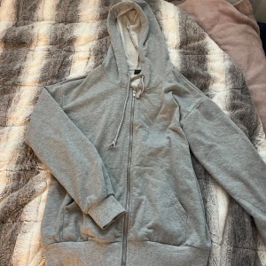 Grå hoodie med dragkedja och huva - Enkel och stilren grå hoodie med dragkedja framtill, huva med snörning och två stora fickor. Tröjan är i mjuk bomullsblandning och har ribbade muddar vid ärmslut och nederkant. Perfekt för chill dagar eller när du vill ha något bekvämt.