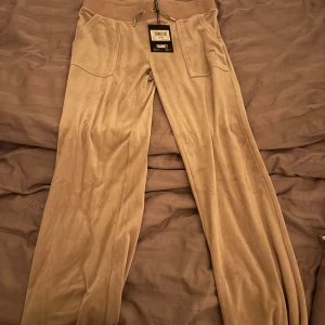 Beiga Juicy storlek S  - Snygga beige velourbyxor i modellen Layla Low Rise Pocketed Flare. Byxorna har låg midja, utsvängda ben, stora fickor fram och bak med knappdetaljer samt bred resår i midjan med snörning.