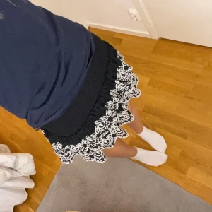 Svart volangkjol med broderi från Lindex - Den är i barn storlek men så pass stretchig att den passar Xs/S. Supersöt svart kortkjol från Lindex med bred smock i midjan och två lager volanger. Kjolen har vita broderade detaljer längs volangerna som ger en unik och lekfull look. Perfekt för dig som gillar att sticka ut lite extra!