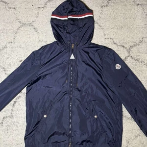  Windbreaker från Moncler - Svart Moncler vindjacka med huva och dragkedja framtill. Jackan har Moncler-logga på ärmen, snörning vid huvan och två fickor med tryckknappar. Skick 10/10 storlek 4 som motsvarar XL