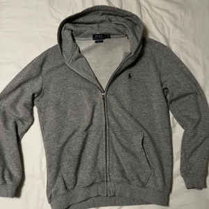 Grå hoodie från Polo Ralph Lauren - Klassisk grå hoodie från Polo Ralph Lauren med dragkedja och huva. Tröjan har långa ärmar, ribbade muddar och två fickor framtill. Broderad marinblå logga på bröstet. Perfekt för chill dagar och enkel att matcha med jeans eller joggers. Säljer den för att den inte längre passar mig så du kan få den för 500 vid snabb affär! 