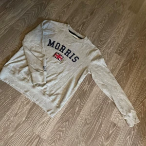 Grå sweatshirt från Morris - Snygg grå sweatshirt från Morris med mörkblå text och brittisk flagga framtill. Tröjan har rund halsringning och långa ärmar, perfekt för en chill och stilren look. Skön bomullskänsla och klassisk passform.