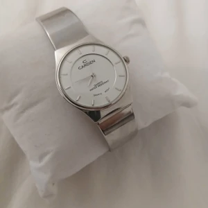 Silverfärgad klocka från Carden - Stilren klocka från Carden med rund vit urtavla och minimalistiska index. Armbandet är i silverfärgat metallnät och boetten är också i silver. Klockan har quartz-urverk och är vattentålig. Perfekt för dig som gillar en clean och klassisk look. Jag vet inte mycket om denna klocka då jag fått ärva den, kom gärna med info om ni vet något om den och kom gärna med prisförslag.
