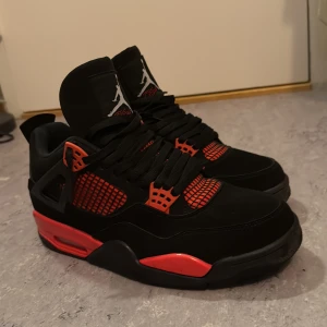 Nike Air Jordan 4 svart/röd sneakers - Säljer ett par Nike Air Jordan 4 sneakers i svart och rött med klassisk Jumpman-logga på hälen. Skorna har meshdetaljer, snörning och en riktigt fet röd sula som poppar. Ovandelen är i mocka och syntet, och insidan är fodrad i rött tyg. Perfekta för dig som vill sticka ut.