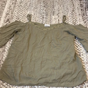 Olivgrön blus med spets från Noisy May - Supersöt olivgrön blus från Noisy May med v-ringning och spetsdetaljer längs halsen. Toppen har offshoulder-stil med smala axelband och vida trekvartsärmar. Storlek S. Köpt från Cassels.