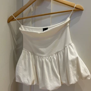 Vit volangig tubtopp Gina Tricot - Supersöt vit tubtopp från Gina Tricot med volang, puffig nederdel. Toppen har en rak, något draperad halsringning och är tillverkad i mjuk bomull. Perfekt för en fräsch och trendig look i sommar.