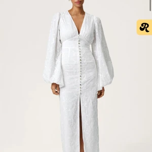 Vit långklänning med puffärmar från Adoore - Superfin vit långklänning med v-ringning, puffiga långärmar och knappar framtill. Klänningen har broderade detaljer och en slits framtill som ger en elegant look. Perfekt för dig som vill sticka ut med en romantisk och trendig stil.