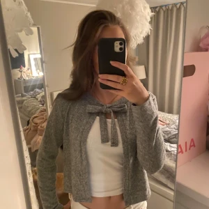 Grå kofta med knyt i halsen - Superfin grå kofta med knytband i halsen. Den är långärmad och har en mjuk, stickad känsla som gör den perfekt att slänga på sig över ett linne eller t-shirt. Enkel och stilren design som passar till många outfits.