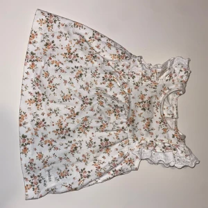 Blommig kortklänning från Newbie - Supersöt vit kortklänning från Newbie med små blommor i rosa, orange och grönt. Klänningen har volangdetaljer vid axlarna, knappar bak och ärmlös design. Perfekt för varma dagar och riktigt gullig stil.