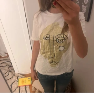 Vit t-shirt med konstnärligt ansikte - Vit t-shirt med korta ärmar och ett stort konstnärligt tryck av ett abstrakt ansikte i guld och svart på framsidan. T-shirten har en klassisk passform och är gjord i mjukt bomullstyg. Perfekt för dig som gillar unik och trendig stil.