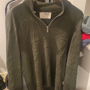 Olivgrön stickad tröja Selected Home - Snygg olivgrön stickad tröja från Selected Homme med strukturerat mönster och half zip-dragkedja vid halsen. Tröjan har lång ärm och ribbad krage, perfekt för lager-på-lager-stil. Passar dig som gillar en modern och avslappnad look.