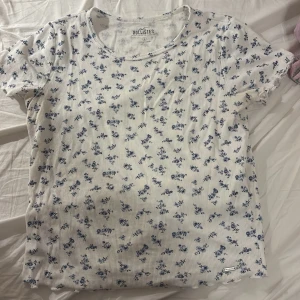 Vit blommig topp från Hollister - Supersöt vit topp från Hollister med blått blommigt mönster. Modellen är ribbad med korta ärmar och rund halsringning. Perfekt för dig som gillar en romantisk och fräsch stil. Materialet är mjukt och stretchigt, vilket gör den riktigt skön att bära. Köpt second hand men är i bra skick! Använd typ en gång för den är lite för stor för mig tyvärr 