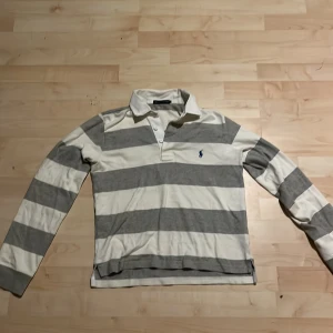 Randig pikétröja från Ralph Lauren - Snygg pikétröja från Ralph Lauren i grått och vitt med breda ränder. Klassisk krage, knäppning med två knappar och lång ärm. Ikoniska loggan broderad på bröstet. Perfekt för en chill och sportig stil. Storleken är för 10-12 åringar har råkat tvätta så den blev förliten 