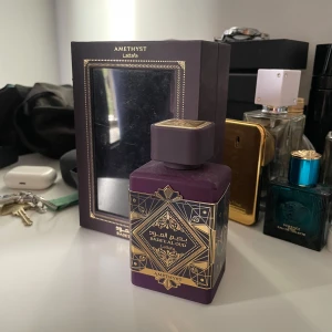 Lattafa Badee Al Oud Amethyst parfym - Lattafa Badee Al Oud Amethyst är en lyxig parfym i en mörklila, fyrkantig flaska med detaljer i guld och orientaliskt mönster. Flaskan har en matchande lila kork och kommer i en elegant ask med spegel. Perfekt för dig som gillar exklusiva dofter och snygg design.