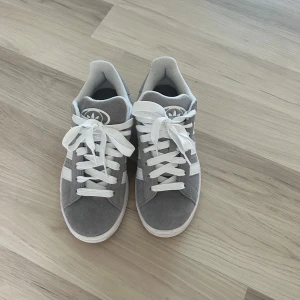 Grå Adidas Campus sneakers  - Snygga Adidas Campus sneakers i grå mocka med klassiska vita ränder och vit sula. Skorna har rund tå, platt sula och vita snören. Perfekta för dig som gillar en clean och tidlös stil med streetkänsla.
