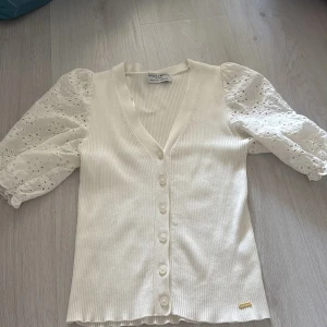 Vit blus med broderade puffärmar från Holly & Whyte - Supersöt vit blus från Holly & Whyte med ribbad kropp och broderade puffärmar i bomull. V-ringad med knappar framtill och fina detaljer på ärmarna. Perfekt för en fräsch och stilren look! 